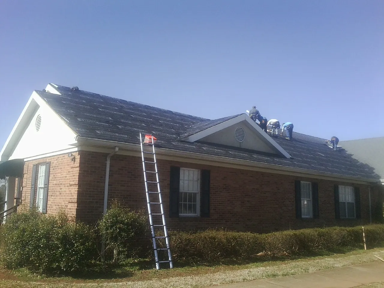 Crew tearing shingles off a brick ranch roof with a ladder set — Metal Roof Repair work by our crew in Colbert